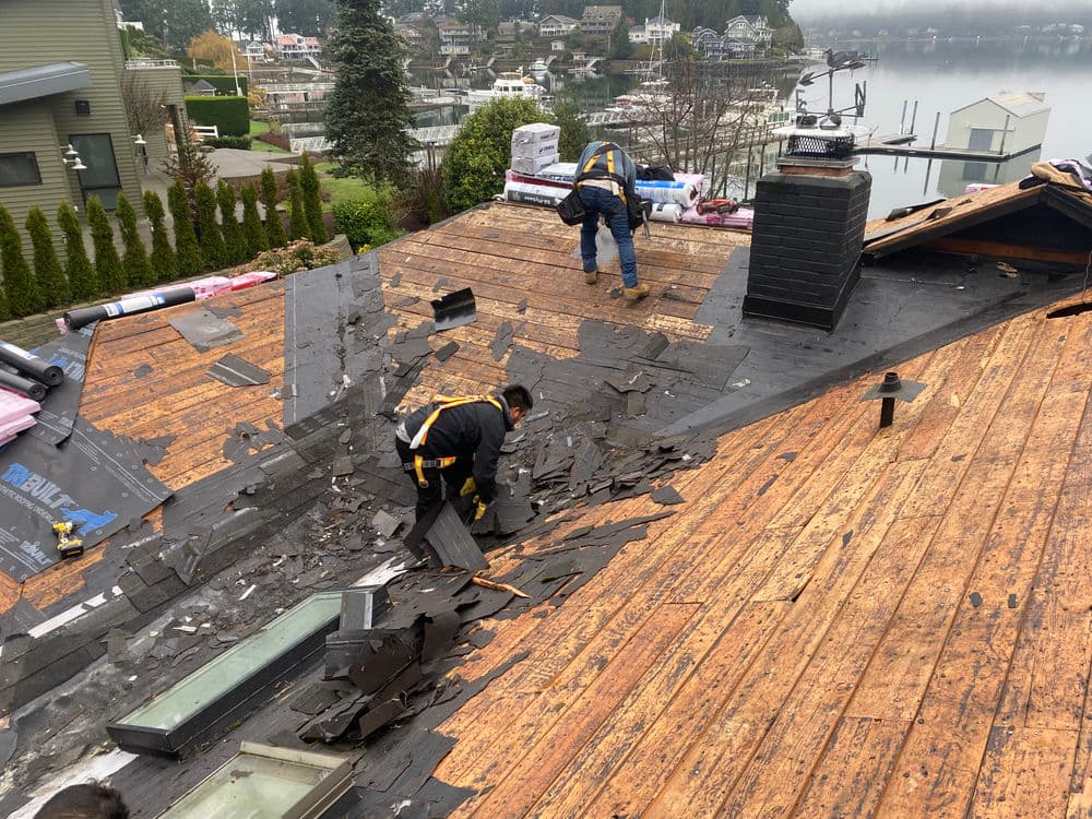 Roofing contractors removing old shingles on a residential rooftop near a lakeside community.